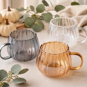 Puede incluir: Tres tazas de cristal con forma acanalada similar a una calabaza. Una taza es transparente, otra es de color ámbar translúcido y la tercera es de color gris oscuro translúcido. Cada taza tiene un cuerpo redondeado y un asa curva lateral.