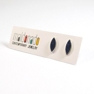 May include: A pair of silver earrings with dark blue oval-shaped stones. The earrings are on a white card with the text "maldonado CONTEMPORARY JEWELRY".