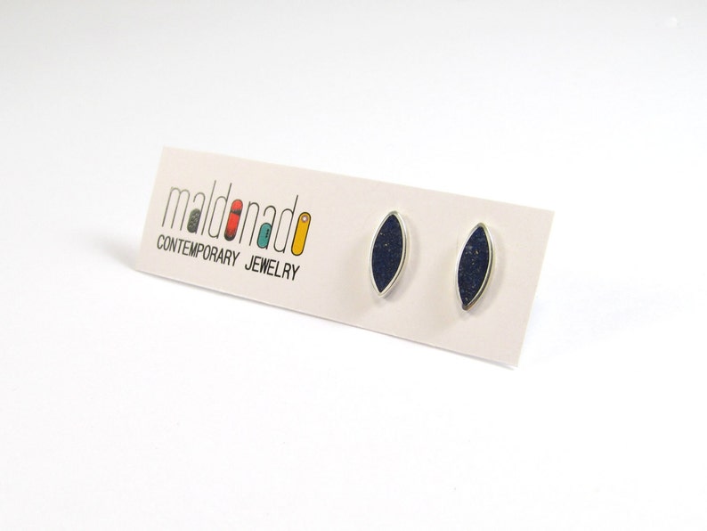 May include: A pair of silver earrings with dark blue oval-shaped stones. The earrings are on a white card with the text "maldonado CONTEMPORARY JEWELRY".
