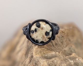 May include: A handmade ring featuring a round, speckled stone set in a dark, wire-wrapped band. The stone has a cream-colored base with black spots. The ring is displayed on a textured, brown surface.