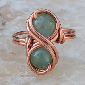 May include: A copper wire wrapped ring with two green stones. The ring is in the shape of an infinity symbol.