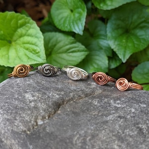 Könnte beinhalten: Fünf Drahtringe mit einem Spiraldesign. Die Ringe sind in verschiedenen Farben: Kupfer, Silber, Schwarz und Gold.