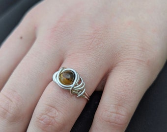 Puede incluir: Un anillo de alambre plateado con una piedra de ojo de tigre marrón.