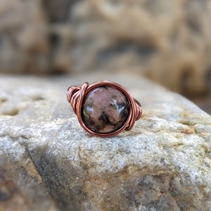May include: A copper wire wrapped ring with a pink and black gemstone. The ring is sitting on a rock.