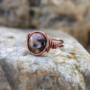 May include: A copper wire wrapped ring with a pink and black gemstone. The ring is sitting on a rock.