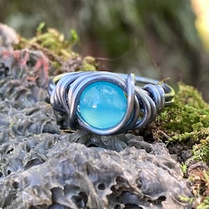 May include: A wire-wrapped ring with a light blue gemstone. The ring is made of silver wire and the gemstone is set in a bezel setting.