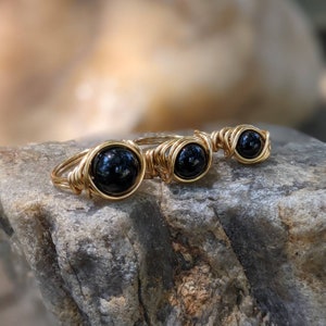 May include: Three gold wire wrapped rings with black gemstone beads. The rings are arranged in order of size, with the largest ring on the left and the smallest ring on the right.