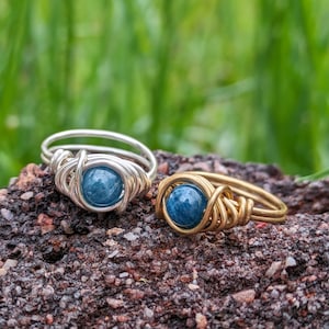 Könnte beinhalten: Zwei drahtgewickelte Ringe mit blauen Edelsteinperlen. Der Ring links ist silber und der Ring rechts ist gold.