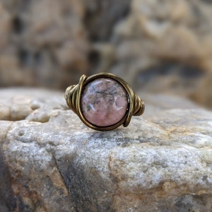 Puede incluir: Un anillo de piedra preciosa rosa y gris con un engaste de alambre de lat&oacute;n. El anillo est&aacute; sobre una roca.