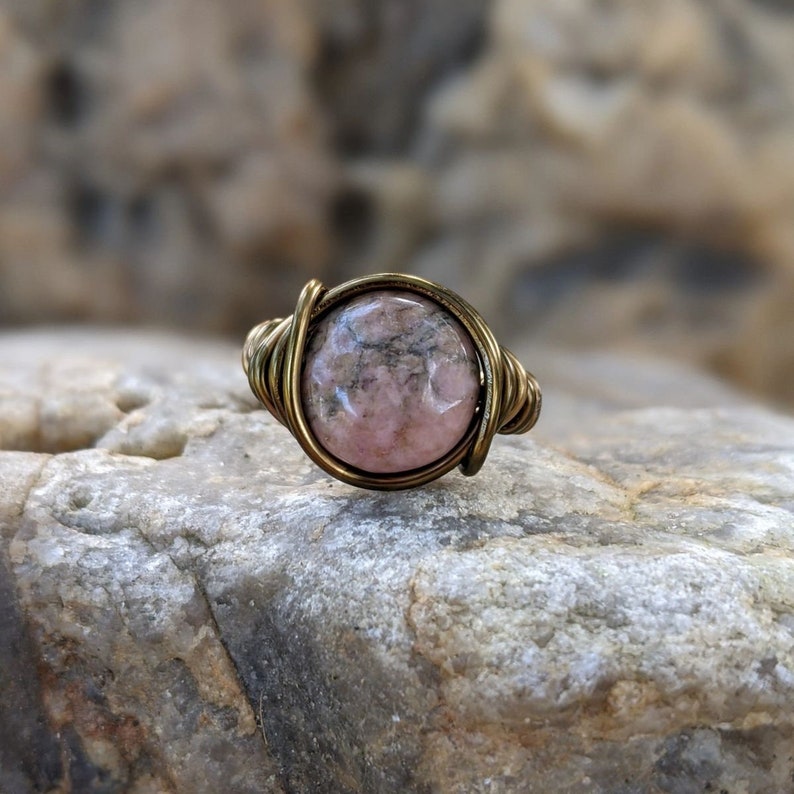 Puede incluir: Un anillo de piedra preciosa rosa y gris con un engaste de alambre de lat&oacute;n. El anillo est&aacute; sobre una roca.