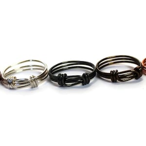 May include: Five wire rings in different colors: copper, silver, black, bronze, and copper. Each ring is made of three strands of wire twisted together with a knot in the center.