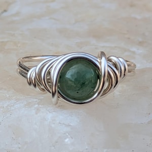 May include: A handcrafted ring featuring a round, green gemstone set in a silver wire wrap design. The ring has a delicate, yet intricate appearance, with the wire carefully coiled around the stone. The band is a simple, double-strand design.