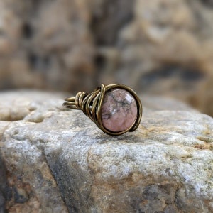 Puede incluir: Un anillo con una piedra rosa y gris envuelta en alambre de lat&oacute;n.