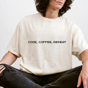 Może przedstawiać: Kremowy t-shirt z czarnym, pikselowanym napisem "CODE, COFFEE, REPEAT". Koszulka ma luźny krój. Osoba nosi również czarne dżinsy z przetarciami.