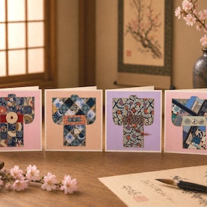 Könnte beinhalten: Vier Grußkarten mit Kimono-Designs. Jede Karte zeigt ein einzigartiges Kimono-Muster in Blau-, Rosa-, Orange- und Lilatönen. Die Karten liegen auf einer Holzoberfläche mit Kirschblüten und einem Kalligraphiepinsel.