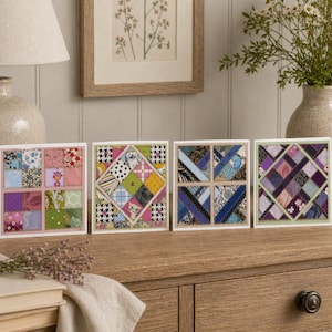 Puede incluir: Cuatro tarjetas de felicitación cuadradas con diseños de patchwork. Cada tarjeta presenta una disposición única de cuadrados de tela en varios colores y patrones, incluyendo motivos florales, geométricos y abstractos. Las tarjetas se exhiben sobre una superficie de madera.