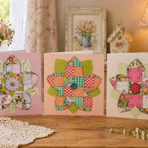 May include: Three handmade greeting cards with floral designs. Each card features a patchwork flower in shades of pink, orange, and cream, with intricate fabric patterns. The cards are displayed on a wooden table, with a vase of pink roses in the background.