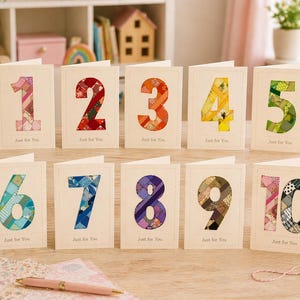 May include: A set of ten greeting cards with numbers 1 through 10, each featuring a patchwork design in various colors. The text "Just for You" is printed below each number. The cards are arranged on a wooden surface.