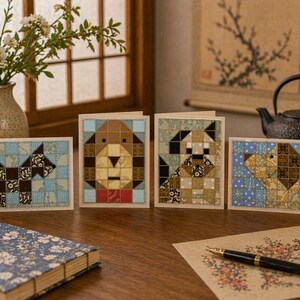 May include: Four greeting cards with patchwork dog designs in various colors, including blue, brown, and tan. The cards are arranged on a wooden surface with a floral notebook, pen, and vase of flowers.