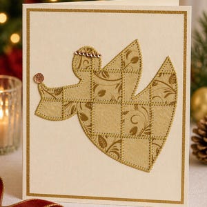 May include: A cream-colored Christmas card featuring a gold angel applique with floral patterns. The angel is adorned with sequins and a gold glitter border. The card is a festive holiday decoration.