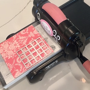 Puede incluir: Una máquina de troquelado negra y rosa con un mango rosa. La máquina tiene una plataforma transparente con un papel estampado rosa y blanco y una matriz de metal en la parte superior. La máquina tiene el texto "Big Shot" impreso en ella.