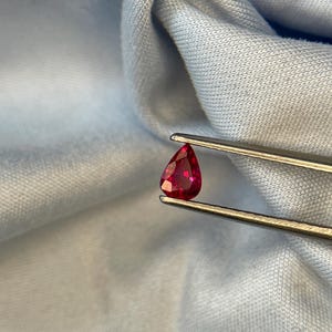 Puede incluir: Una gema roja vibrante, en forma de pera, es sostenida por unas pinzas contra un fondo de tela azul claro. Las facetas de la gema reflejan la luz, mostrando su claridad y rico color. Para la fabricación de joyas.