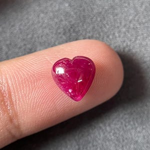 Puede incluir: Una gema de rubí pulida en forma de corazón. La piedra roja oscura se sostiene entre dos dedos. El rubí refleja la luz, resaltando su superficie lisa. El fondo es gris liso.