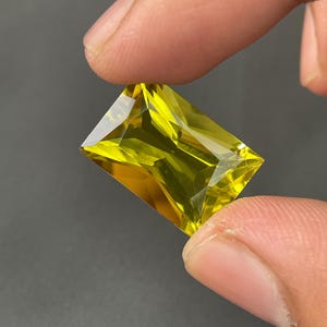 Può includere: Una gemma rettangolare e sfaccettata di un vivace colore giallo. La gemma è tenuta tra due dita, mostrando la sua chiarezza e brillantezza. Il taglio della pietra crea molteplici superfici riflettenti.