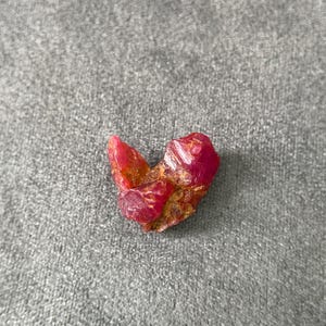 May include: A raw, unpolished gemstone with a vibrant deep pink and red coloration. The stone features an irregular, jagged shape with visible crystalline textures and earthy, brownish-gold mineral inclusions embedded within the crevices of the surface.