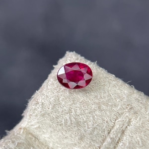 May include: A faceted, oval-cut ruby gemstone. The gem is a deep red colour, with visible internal structures. The ruby is displayed on a white, textured surface against a dark grey background. The gemstone is a focal point.