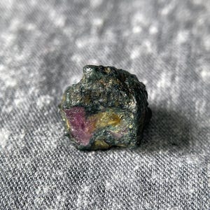 Könnte beinhalten: Ein roher, ungeschliffener Edelstein mit dunkelgrüner Außenseite und sichtbaren rosa und gelben Flecken. Der Stein hat ein raues, natürliches Aussehen, was darauf hindeutet, dass er sich in seinem Rohzustand befindet. Die Farben sind lebendig.