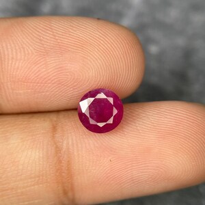 May include: A round, faceted ruby gemstone. The gem is a deep, vibrant red colour, with multiple facets creating a starburst pattern. The ruby is held between two fingers against a neutral background.