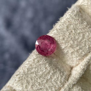 Puede incluir: Una gema de rubí tallada en forma redonda y facetada. La gema es de un color rojo rosado intenso y está colocada sobre una tela crema texturizada. El fondo es un azul grisáceo oscuro borroso.