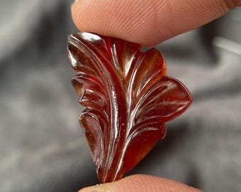 May include: A carved, reddish-brown gemstone shaped like a leaf. The gemstone is held between two fingers, showcasing its intricate details and polished surface. The leaf design features detailed veining.