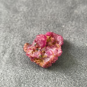 Può includere: Un esemplare di cristallo di rubino grezzo e non lucidato che presenta un ammasso di formazioni esagonali color magenta vibrante e rosa intenso. Il minerale mostra una superficie ruvida e strutturata con materiale di matrice marrone terroso.