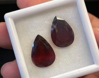 May include: Two faceted, teardrop-shaped, deep red gemstones are displayed in a white square container. The gems have a polished, reflective surface, and are set against a white, textured background. The container is held in a person's hand.