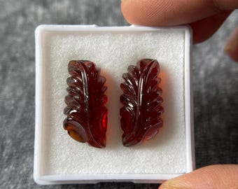 May include: Two carved, reddish-brown gemstone pendants in a white box. Each pendant is leaf-shaped with intricate detailing. The gemstones are held by a person's hand, showcasing their color and design. Ideal for jewelry making.