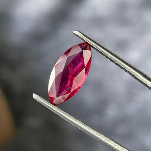 Puede incluir: Una gema de rubí facetada, de forma ovalada. El rubí es de color rojo intenso y está sujeto por unas pinzas plateadas sobre un fondo gris borroso. Las facetas de la gema reflejan la luz, creando un efecto brillante.