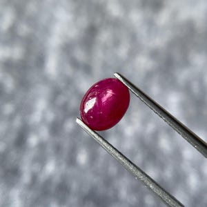 Könnte beinhalten: Nahaufnahme eines ovalen Rubins in tiefroter Farbe, gehalten von einer silbernen Pinzette. Der polierte Edelstein hat eine glatte Oberfläche. Der Hintergrund ist unscharf. Schmuck.