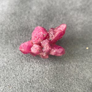 Op de afbeelding: Een cluster van ruwe, ongeslepen robijnkristallen. De kristallen hebben een diepe, levendige roze-rode kleur, met een ruw, getextureerd oppervlak. De cluster is klein en de kristallen zijn dicht bij elkaar gegroepeerd.