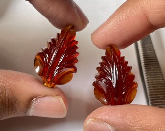 May include: Two carved amber-colored leaf-shaped gemstones are held between fingers. The gemstones have intricate details, with a reddish-brown hue and a glossy finish. The background is a light color, with a measuring tool visible.