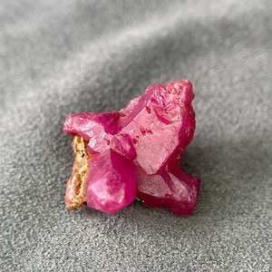 May include: A raw, uncut ruby gemstone with a vibrant pink hue. The crystalline structure of the ruby is visible, with sharp edges and a rough texture. The gemstone is set against a gray background.