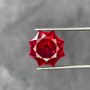 May include: A faceted, star-shaped red gemstone held by tweezers against a gray background. The gem has a complex cut with multiple facets, reflecting light. The color is a deep, vibrant red, and the gem appears to be of high quality.