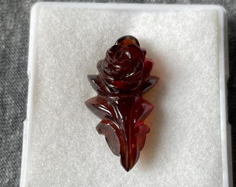May include: A carved, dark red rose pendant displayed in a white plastic case. The rose is intricately detailed, with visible petals and leaves. The pendant is made of a translucent material, possibly a gemstone or resin.