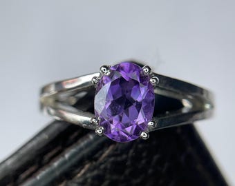 May include: A silver ring featuring a large, oval-cut purple gemstone. The gemstone is set in a split-shank band with small prongs securing the stone. The ring is displayed against a neutral background.