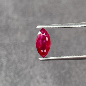 May include: A marquise-cut gemstone with a vibrant, deep red color and faceted surface. The stone is held securely by a pair of silver-toned metal tweezers, showcasing its elongated, pointed shape and reflective clarity in a clear, focused view.