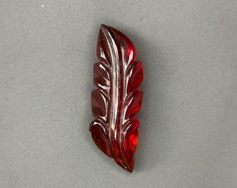 May include: A carved, translucent, red feather-shaped object. The feather has detailed veining and a glossy finish. The object is set against a neutral gray background, highlighting its intricate design and color.