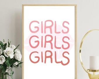 May include: A framed print with the word "GIRLS" repeated three times in pink, with a pink lip-shaped design in the background. The print is in a gold frame and is displayed on a white surface. A bouquet of white roses and a gold candle holder are also visible.