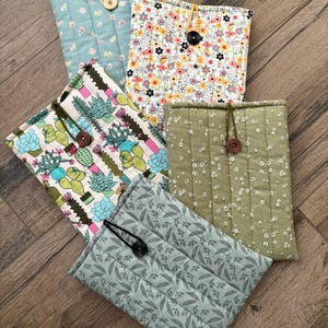 May include: A collection of quilted fabric book covers in various floral and botanical patterns. The covers feature button closures and include designs with cacti, small flowers, and leaf patterns. The color palette includes light blue, green, and white.