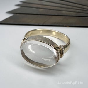 Op de afbeelding: Een zilveren ring met een grote, ovale, heldere edelsteen. De ring heeft een eenvoudige band en een bezel setting. De edelsteen is glad en gepolijst en reflecteert licht. De ring ligt op een witte ondergrond.